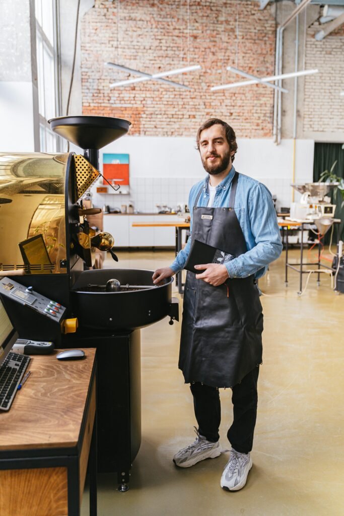 Bearded man with apron using coffee roaster machine indoors, adding notes.